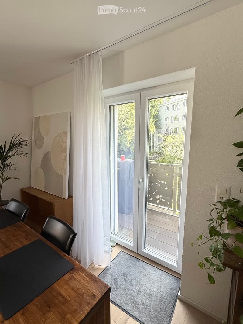 Bright room with large sliding glass doors leading to a balcony, wooden table with chairs, plants, and artwork on the wall.