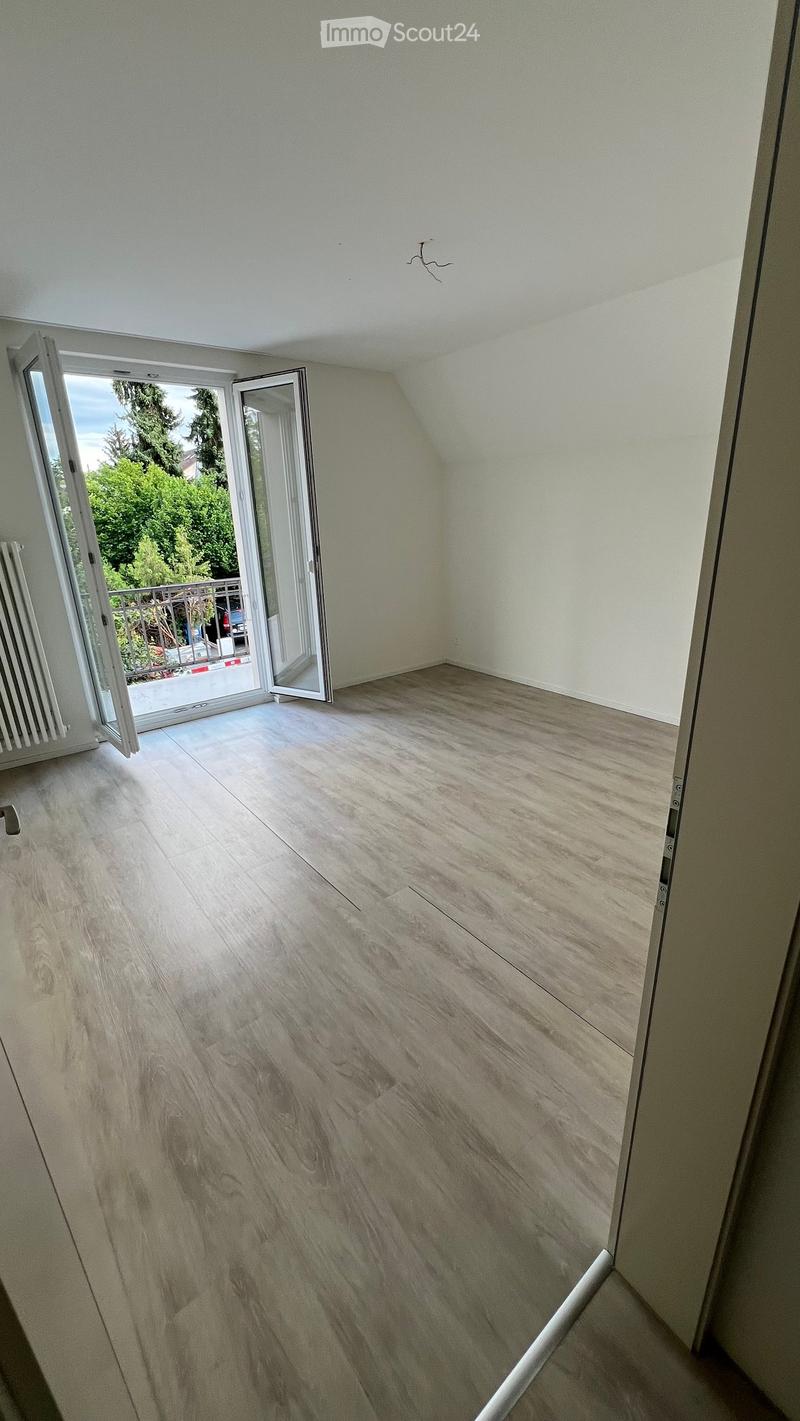 Empty room with light wooden flooring and white walls, featuring an open sliding door leading to a balcony area with outdoor greenery visible.