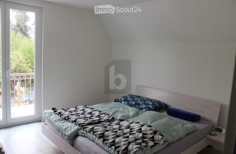 Bedroom with a single bed, pillows, and a blanket. Large window with a view of greenery and a balcony.