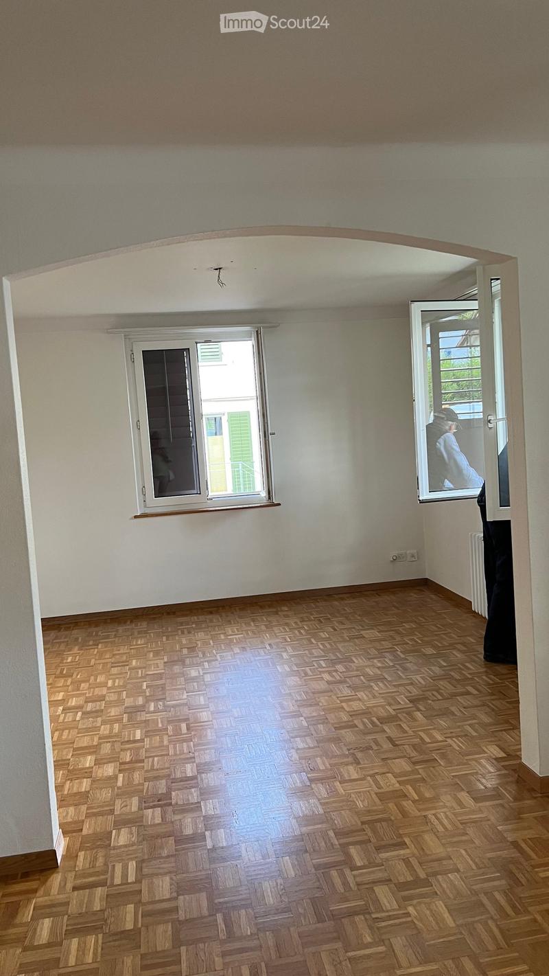 Empty room with wooden herringbone flooring, white walls, and two windows with white frames. The room has a simple, clean design with no furniture or decorations.