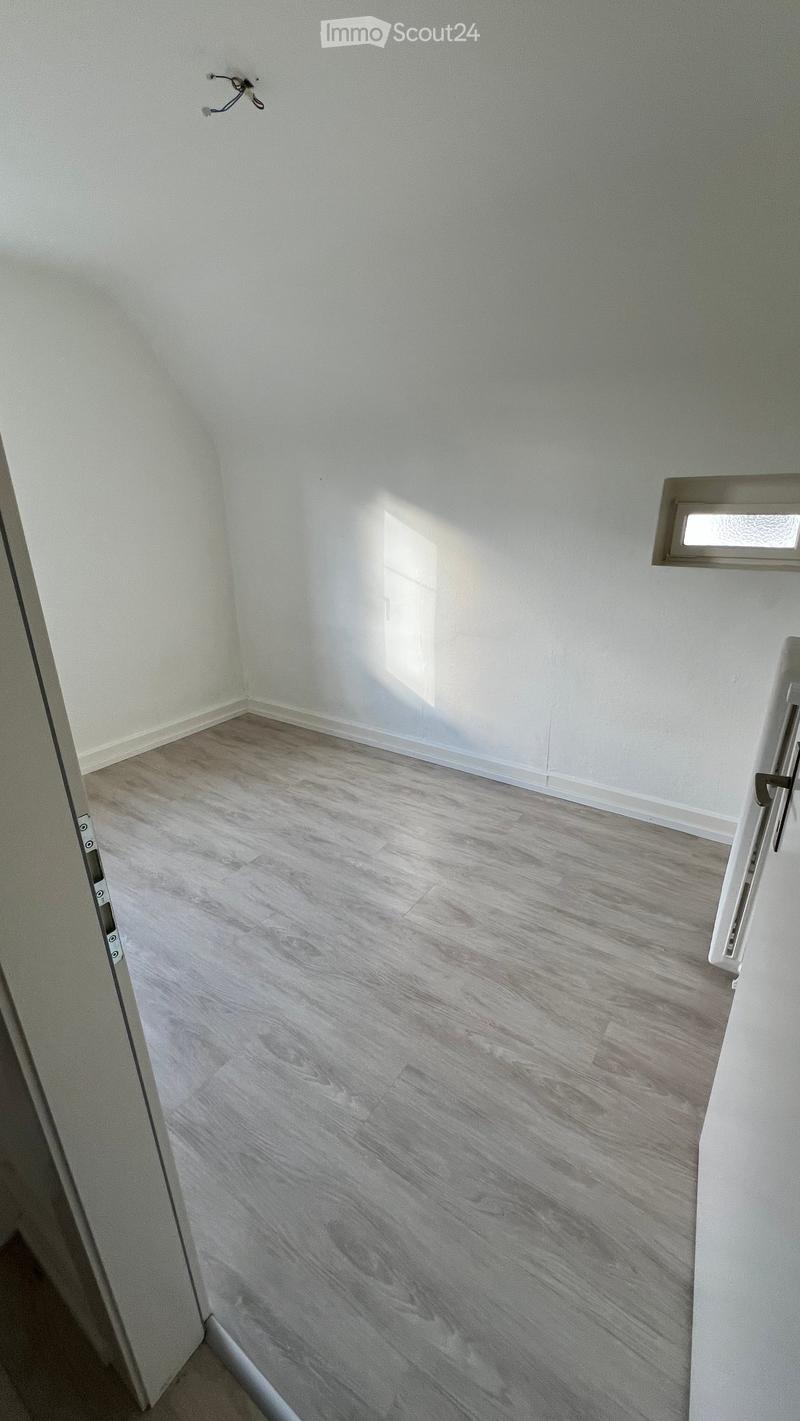 Empty room with light grey wooden flooring and white walls, featuring a small window and a partially visible door frame.