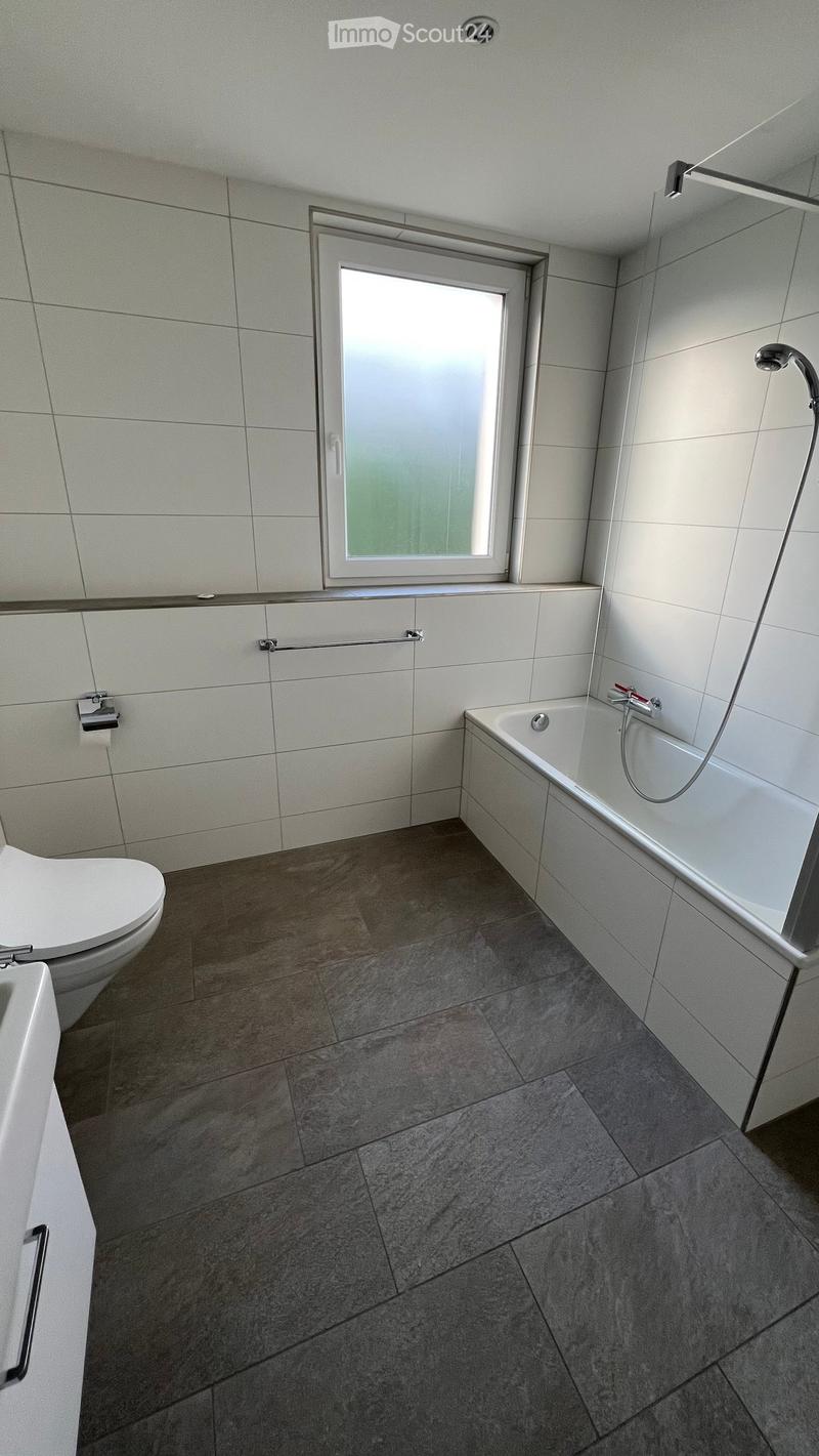 Modern bathroom with a bathtub, toilet, and a window above the bathtub. The walls are tiled, and the floor is made of large, dark tiles.