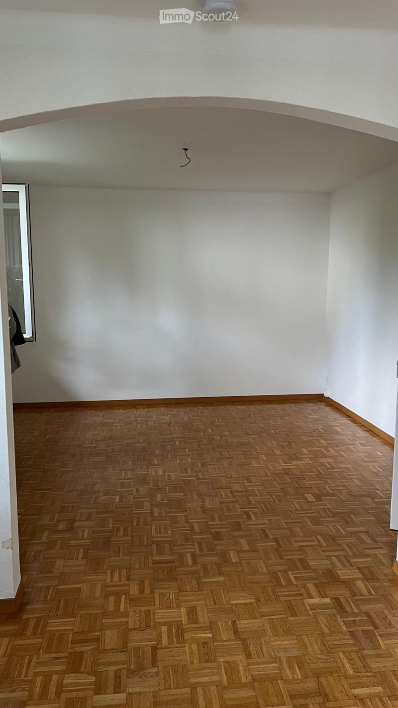 Empty room with wooden herringbone flooring and white walls, featuring a visible smoke detector and a partially visible mirror in the adjacent space.