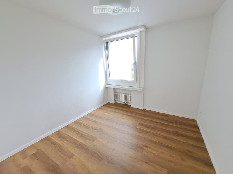 Empty room with wooden flooring, white walls, a window with white frame, and a radiator below the window.