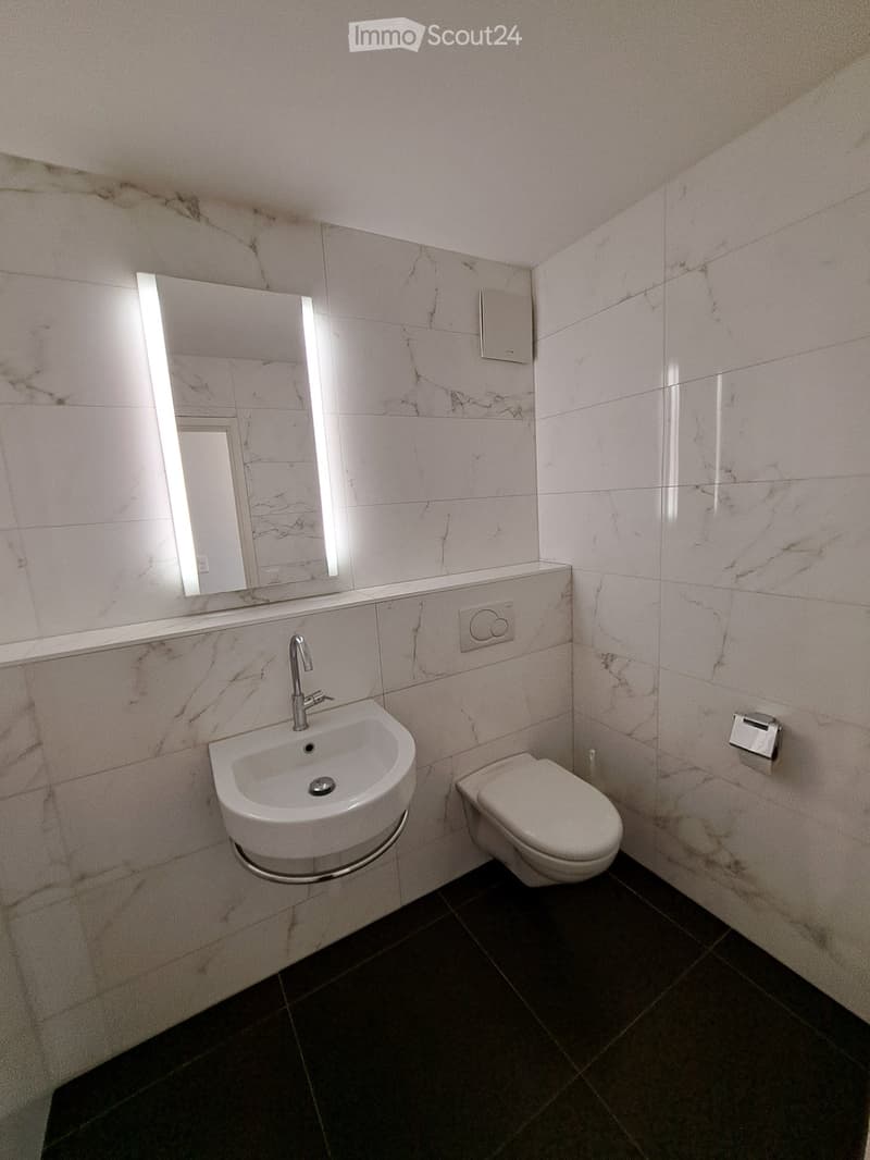 Modern bathroom with marble tiles, a white sink with a chrome faucet, a toilet, and a large illuminated mirror above the sink.