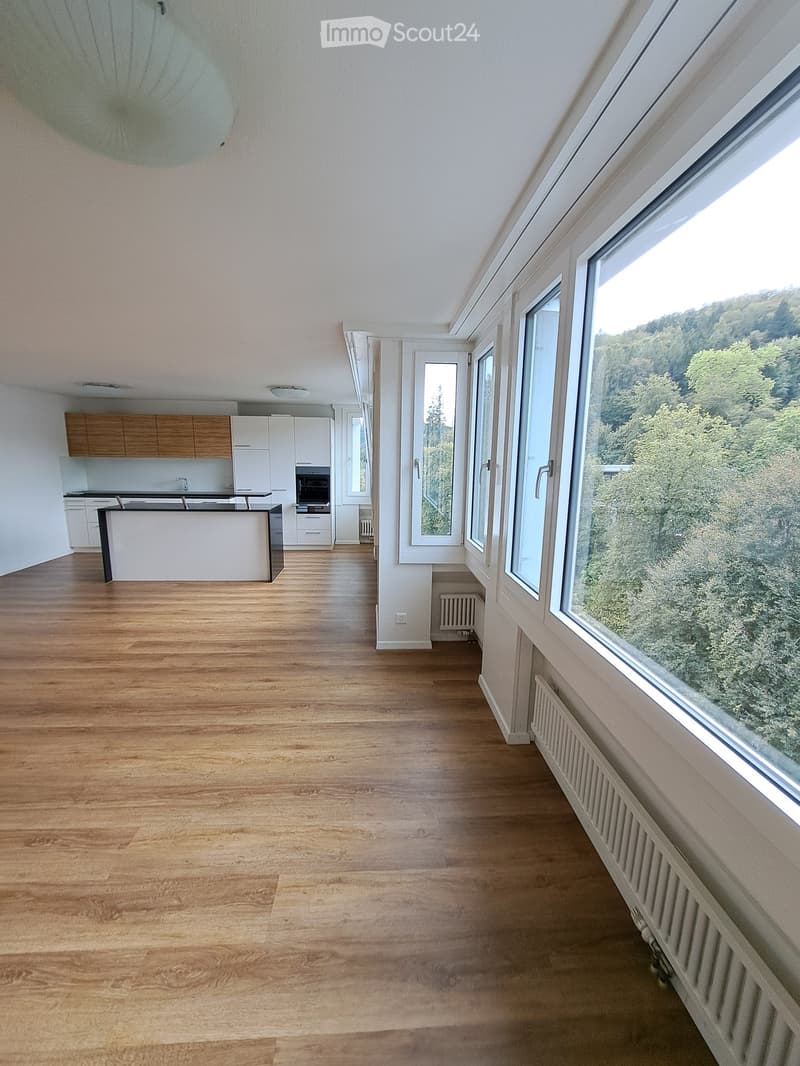 Open-plan living and kitchen area with wooden flooring, large windows overlooking greenery, modern kitchen with island, cabinets, and appliances.