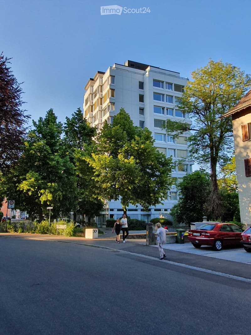 A multi-story residential building with large windows and balconies, surrounded by trees and greenery. A few people are walking on the sidewalk in front of the building, and a red car is parked nearby.
