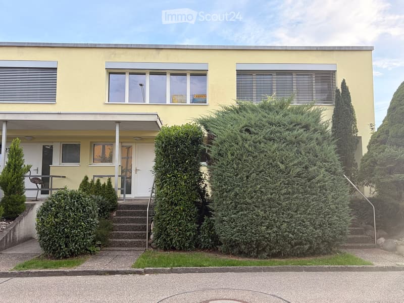 Two-story yellow house with white trim, multiple windows, a small porch, and a landscaped garden with trimmed bushes and trees.