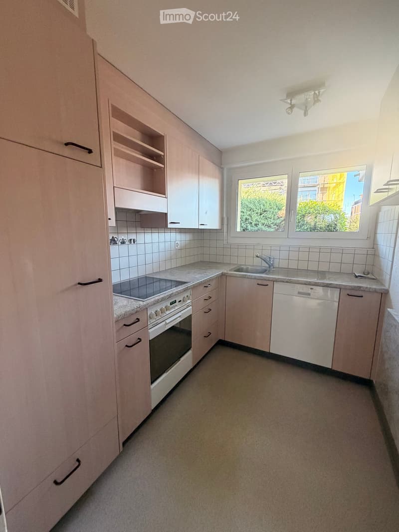 Modern kitchen with white cabinets, granite countertops, built-in oven, sink, and dishwasher. The space is well-lit with natural light from a window overlooking greenery.