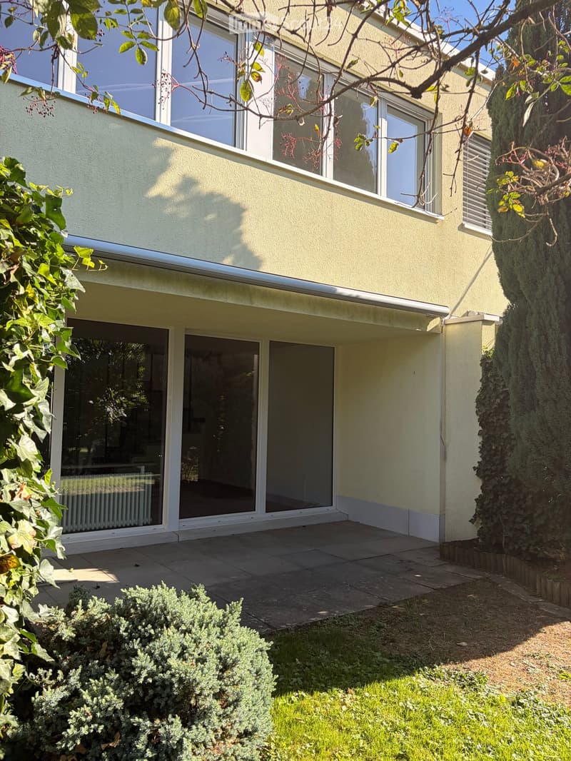 Two-story house with large windows, sliding glass doors, and a well-maintained garden with shrubs and grass.