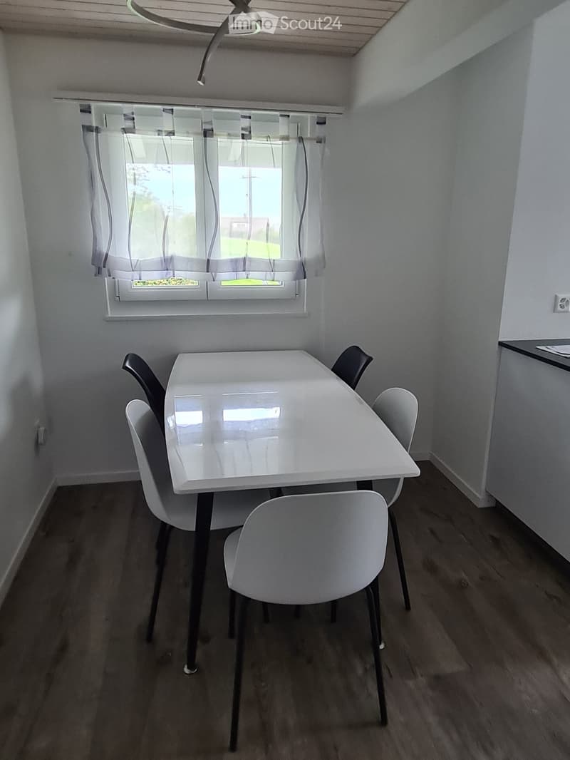 Dining area with a white table and four chairs, natural light from a window with sheer curtains, wooden flooring.