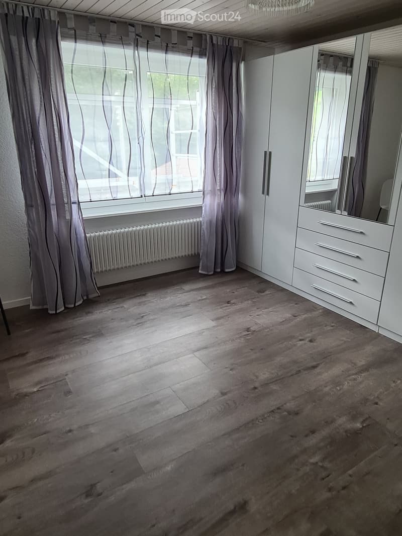 Empty bedroom with wooden flooring, white built-in wardrobe, large window with grey curtains, radiator, and a view of the outside.