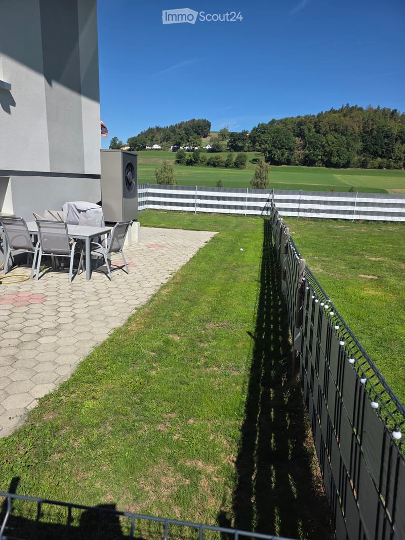 A well-maintained garden with a paved patio area featuring a table and chairs, surrounded by a white fence and lush green grass, with a scenic countryside view in the background.