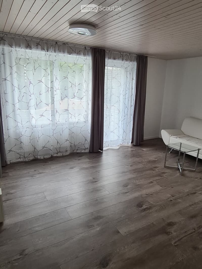 Bright living room with wooden flooring, large windows with patterned curtains, a white sofa, and a glass coffee table.