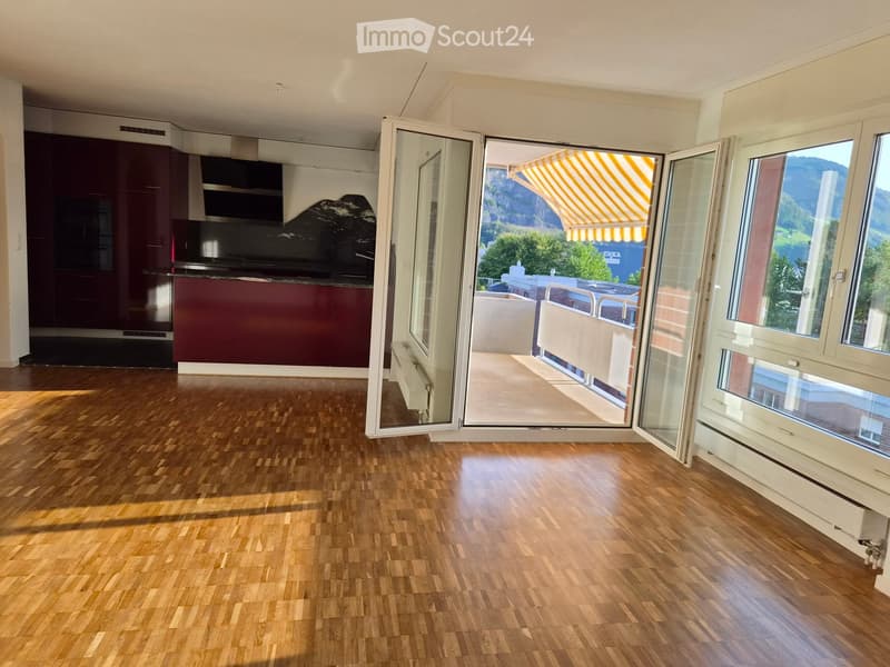 Empty room with wooden flooring, large open sliding doors leading to a balcony with a scenic mountain view, and a modern kitchen area visible in the background.