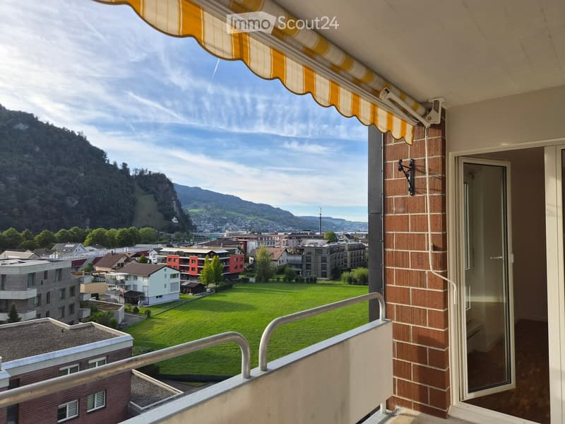 Balcony with a retractable awning, overlooking a cityscape and green fields with distant mountains.