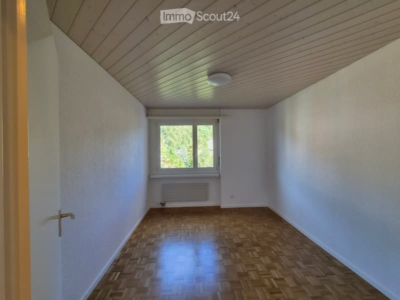 Empty room with wooden parquet flooring, white walls, and a window with a view of greenery. The room features a radiator and a ceiling light fixture.