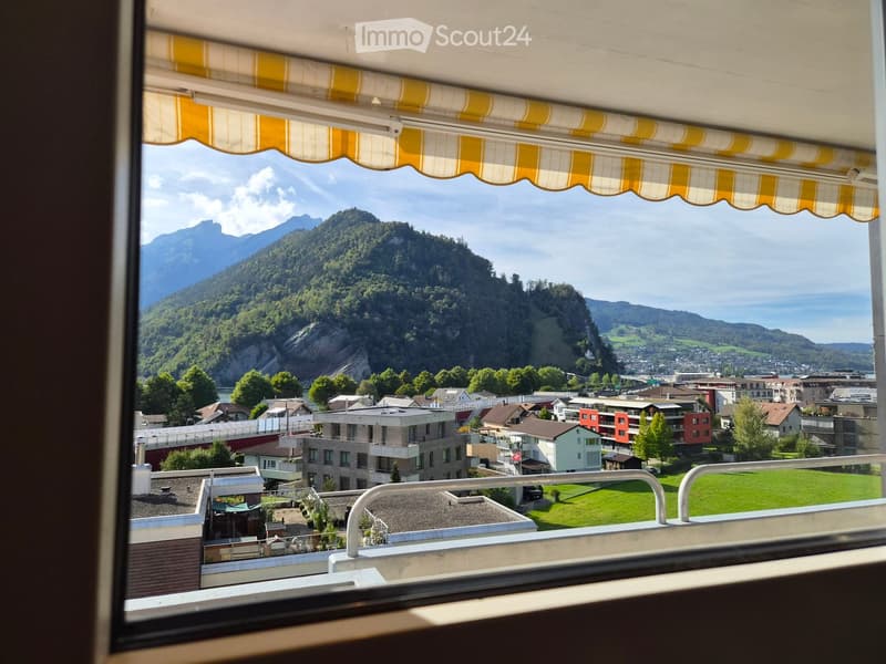 Balcony with a yellow and white striped awning, overlooking a town with houses, green spaces, and distant mountains.