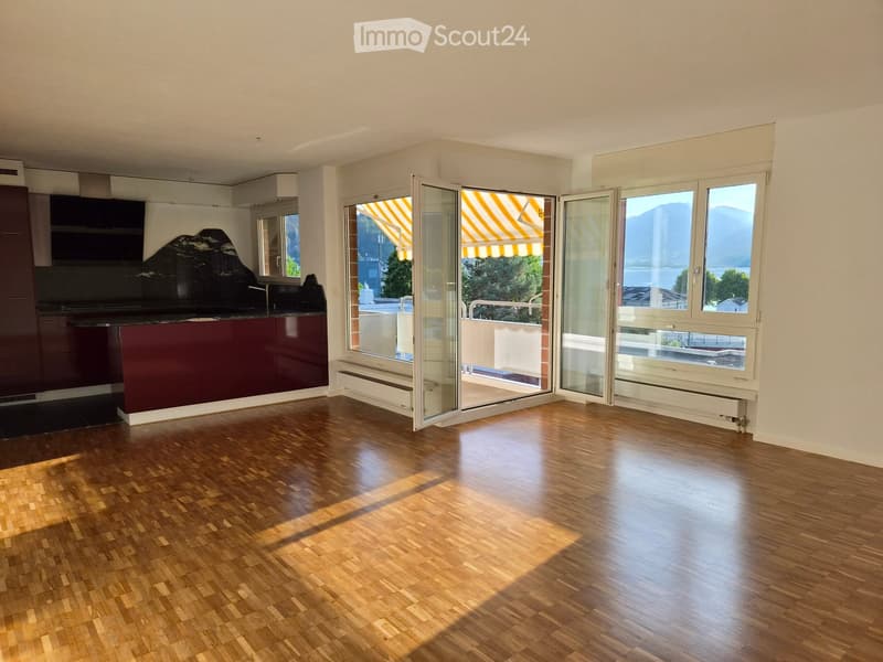 Bright, unfurnished room with wooden flooring, large windows, and a view of mountains and a lake. The room features a modern kitchen area with dark cabinets and a countertop, and large glass doors open to a balcony with striped awnings.
