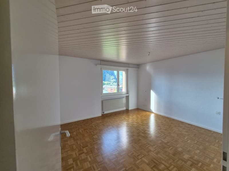 Empty room with wooden flooring, white walls, and a window offering a view of the outside. The room features a radiator below the window and a simple wooden ceiling.