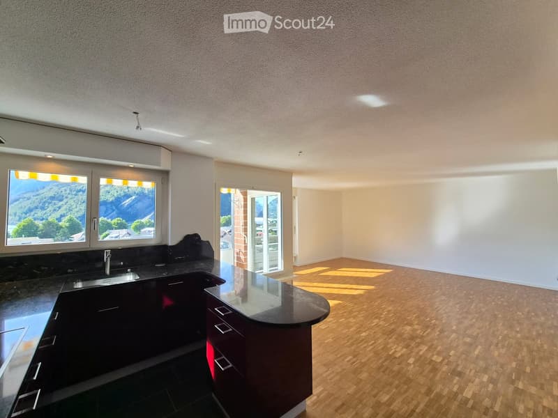 Empty kitchen and living area with dark countertops, wooden flooring, and large windows offering a scenic view.