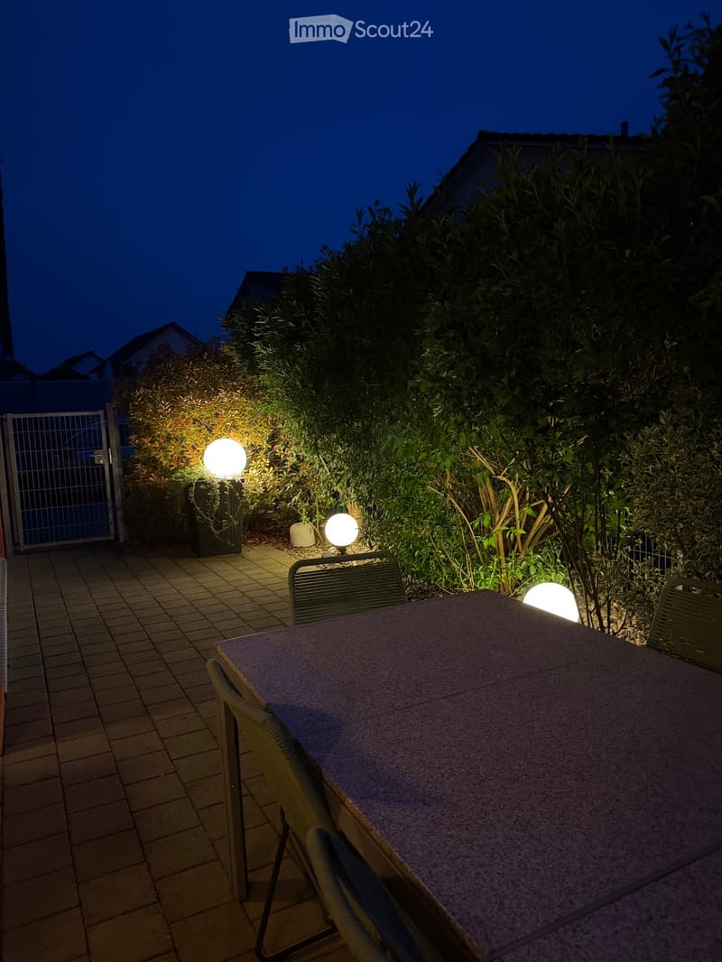 Nighttime outdoor patio with a table, chairs, and spherical lights surrounded by greenery and a gated entrance.
