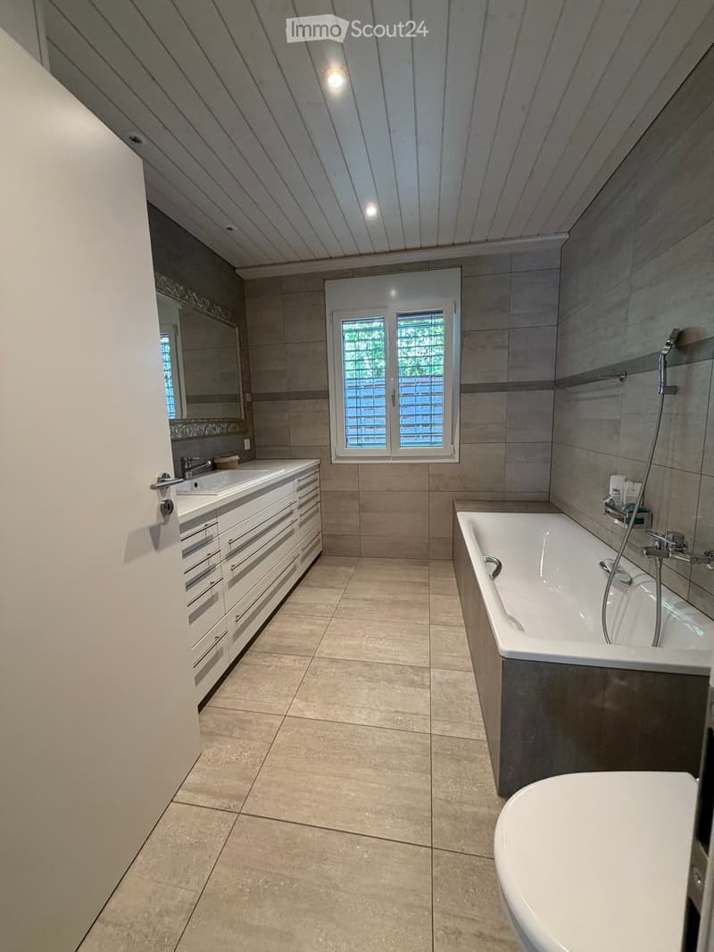 Modern bathroom with a large bathtub, double sink vanity, and a large window with blinds. The space features light-colored tiles and a sleek, contemporary design.