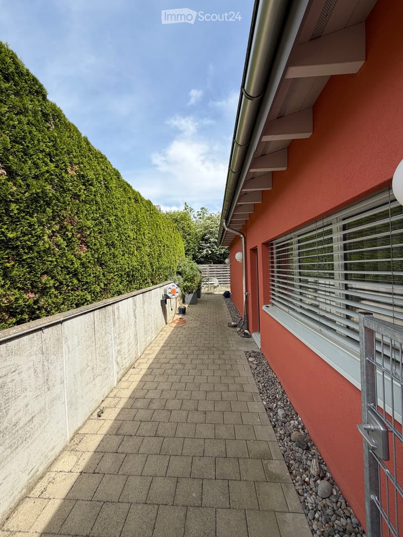 Narrow brick-paved balcony with a low concrete wall on one side, a red building wall on the other, and a small garden area visible in the distance.