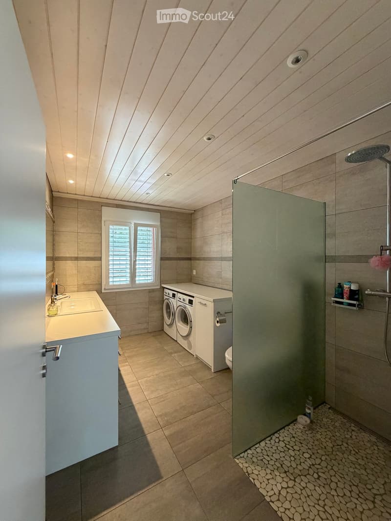 Modern bathroom with a large window, a sink with a faucet, a washing machine and dryer unit, a glass-enclosed shower with a showerhead, and a toilet. The space features beige tiles and a sleek design.