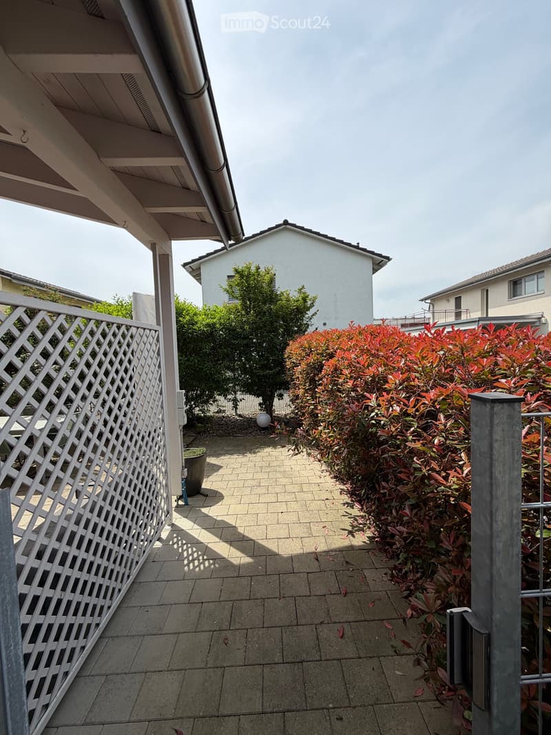 A small, paved outdoor area with a white lattice fence, surrounded by red-leaved shrubs and a tree, with neighboring houses visible in the background.