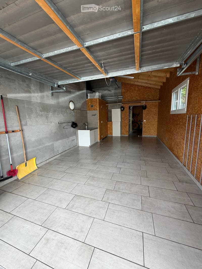 Empty garage with tiled floor, concrete walls, wooden ceiling beams, and a small sink area. Cleaning tools are visible on the left side.