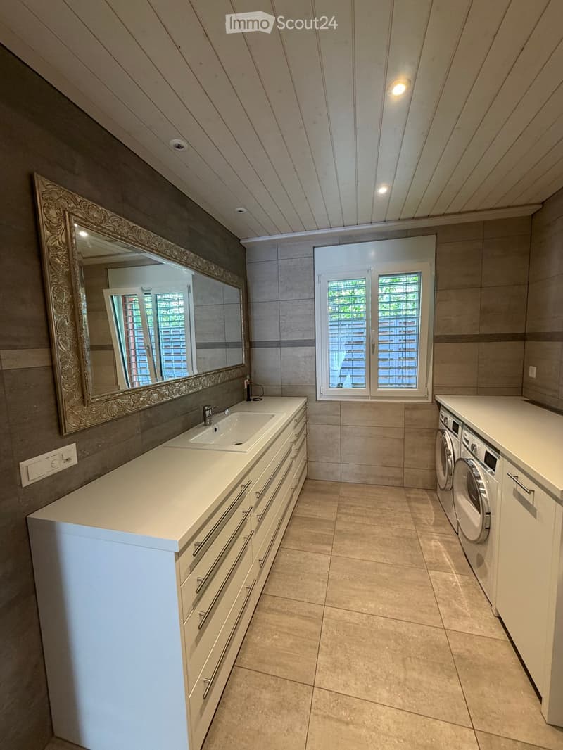 Modern bathroom with a double sink, large mirror, and built-in washing machine and dryer. Features light-colored tiles and a sleek design.