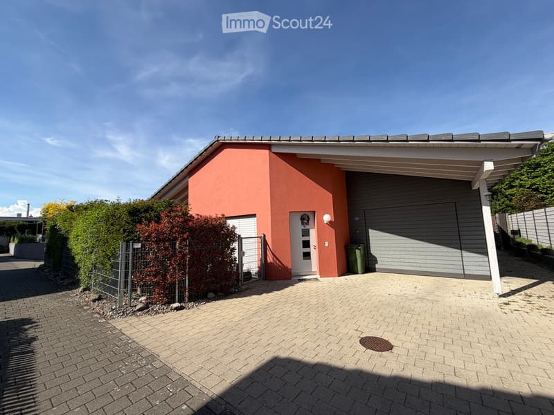 Single-story house with a red facade, white garage door, and a covered entrance area. The driveway is paved with bricks, and there is a small garden area with shrubs and a fence.