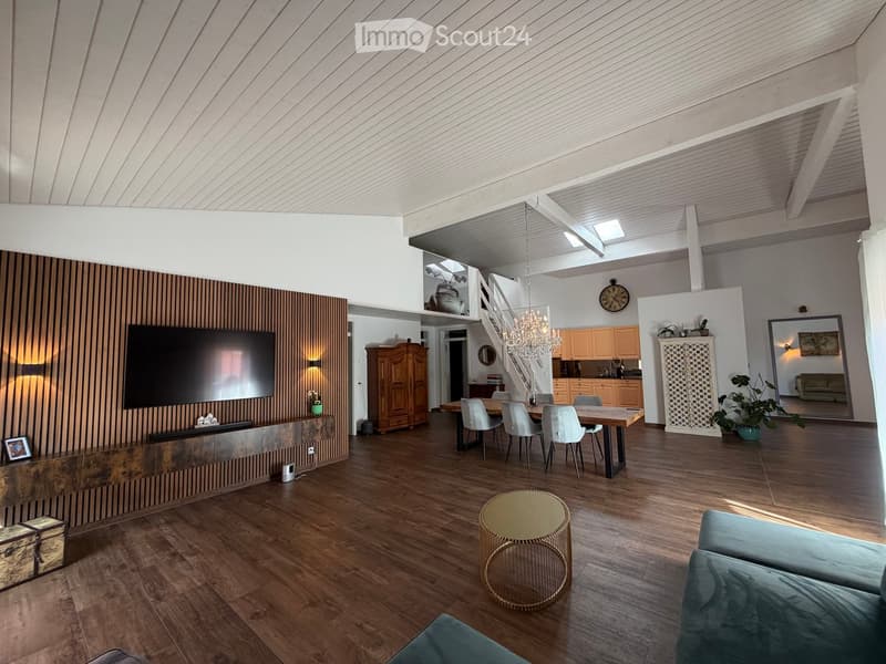 Open-concept living and dining area with wooden flooring, a large TV on a wooden wall, a dining table with chairs, a sofa, a coffee table, and a chandelier hanging from the ceiling.