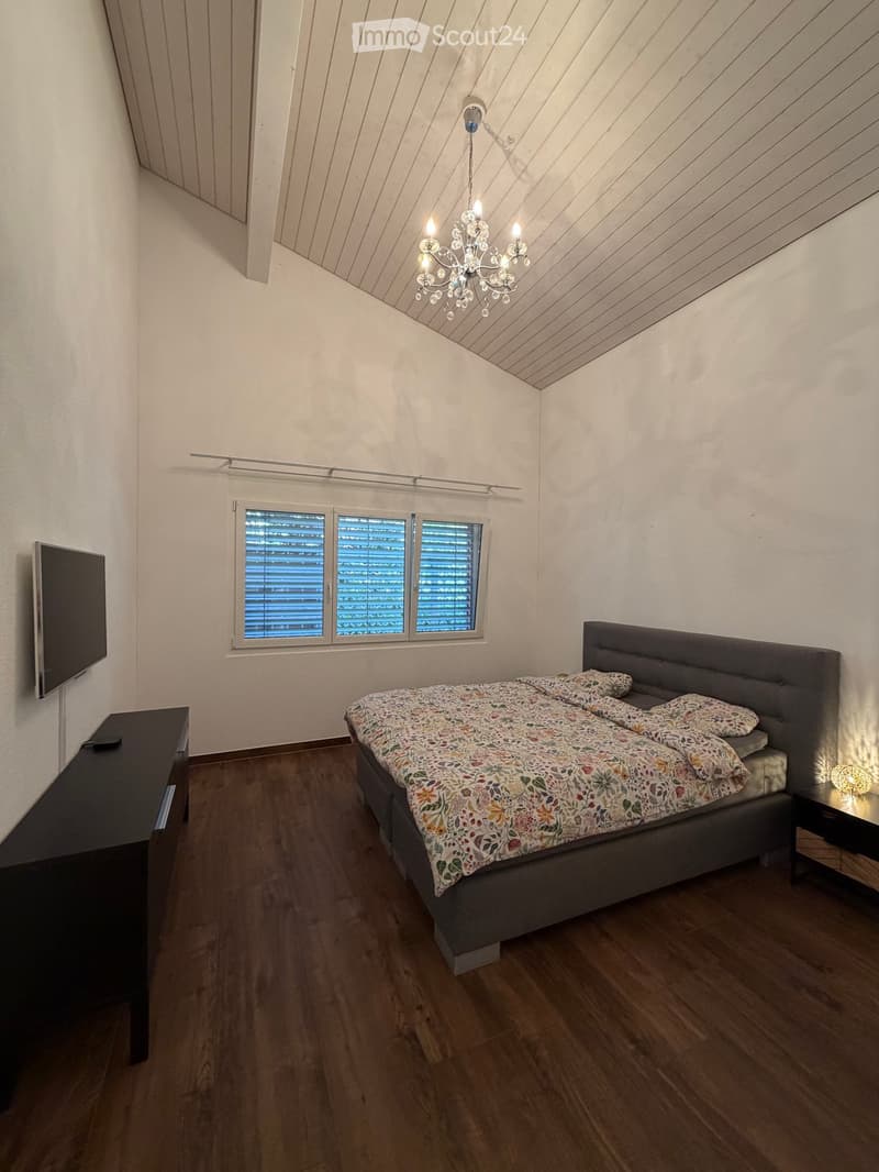 A modern bedroom with wooden flooring, a large bed with a floral duvet, a nightstand with a lamp, a TV on a stand, and a chandelier hanging from the sloped ceiling.