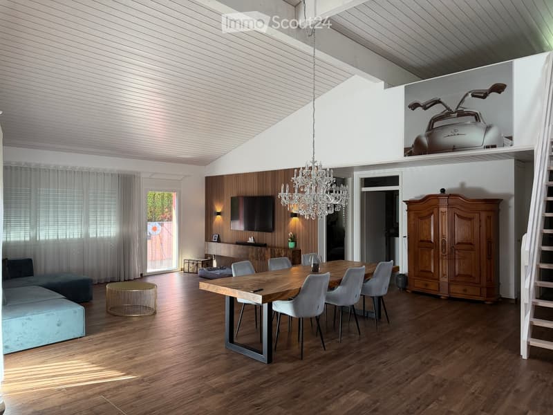 Modern living and dining area with wooden flooring, a large chandelier, a sofa, ottoman, dining table with chairs, and a wooden cabinet.