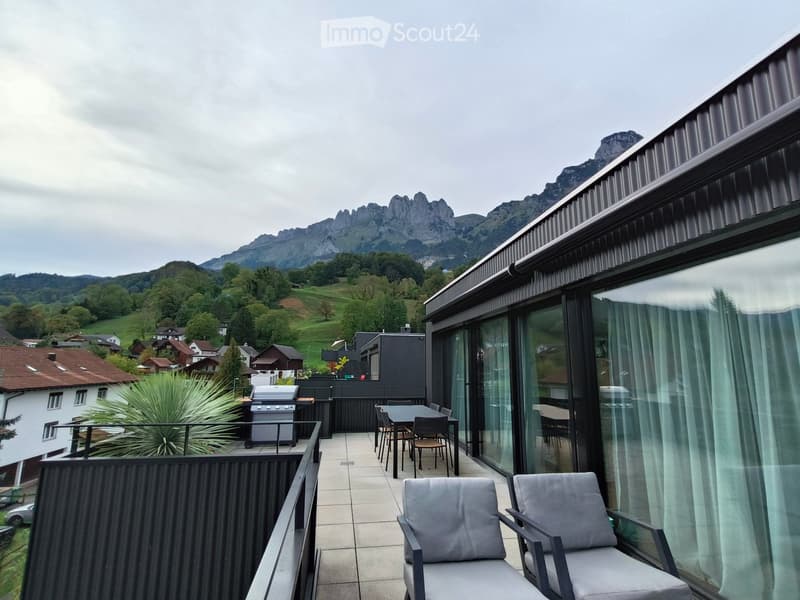 Balcony with modern furniture, overlooking a scenic mountain range and surrounding greenery.