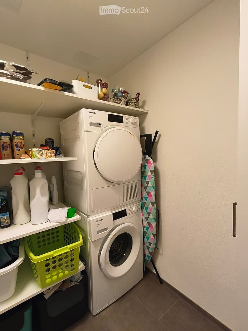 A small laundry area with a white washing machine and dryer stacked, shelves with cleaning supplies, and a drying rack.