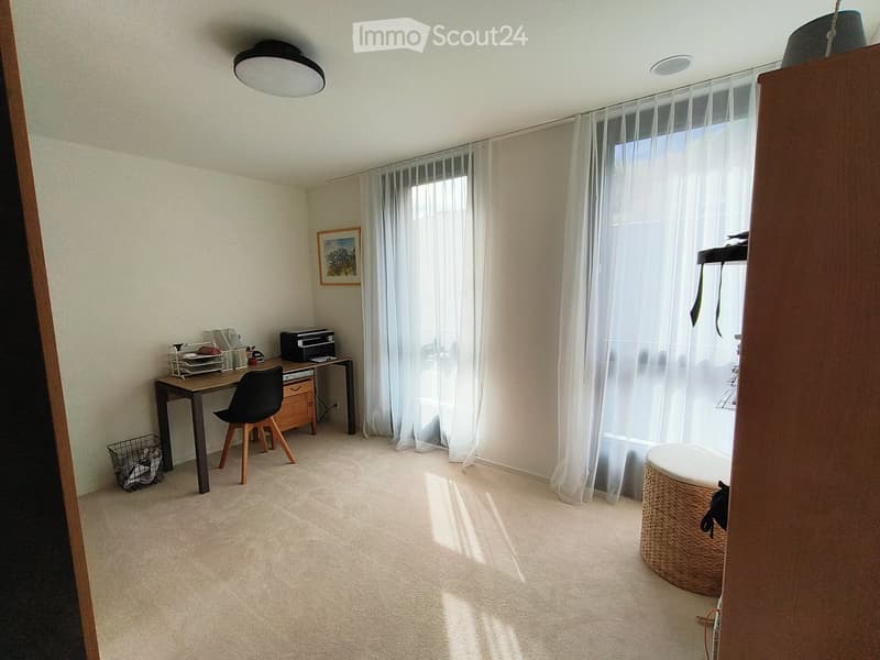 Bright, carpeted room with a desk, chair, printer, and a wicker stool near a large window with sheer curtains. A picture frame hangs on the wall, and a small basket sits on the floor.