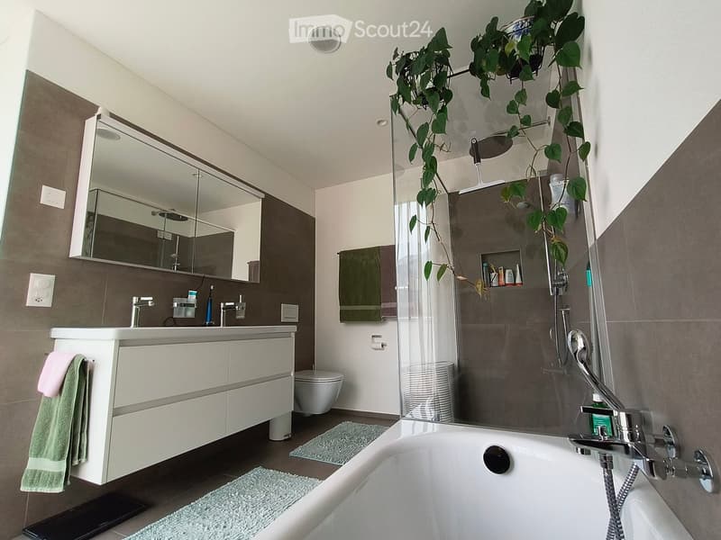 Modern bathroom with a bathtub, sink, toilet, large mirror, and a glass-enclosed shower area. Features include a white vanity with storage, green towels, and a potted plant near the shower.