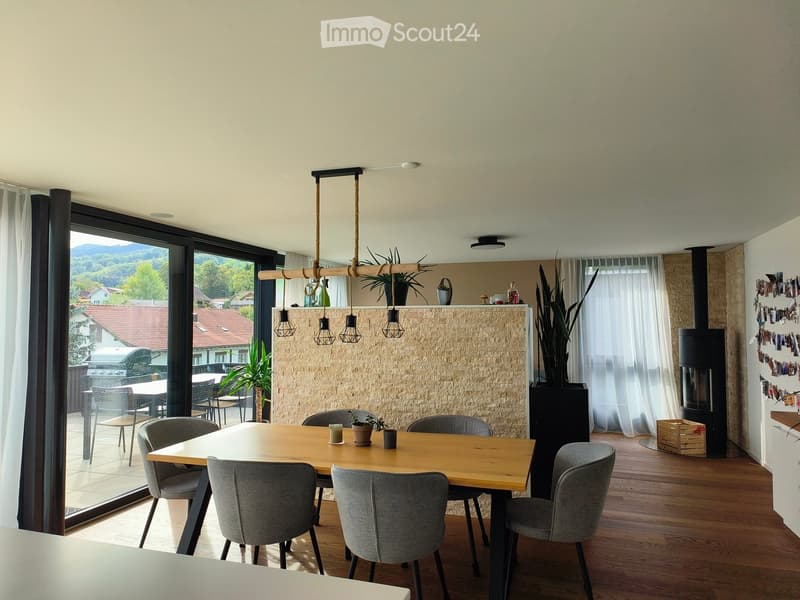 Modern dining area with a wooden table, grey chairs, pendant lights, and a view of the outdoor patio. Features a stone fireplace and large windows with curtains.