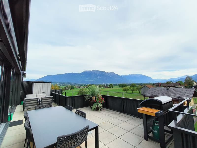 Balcony with a table, chairs, barbecue, and potted plants, overlooking a scenic valley and mountains.