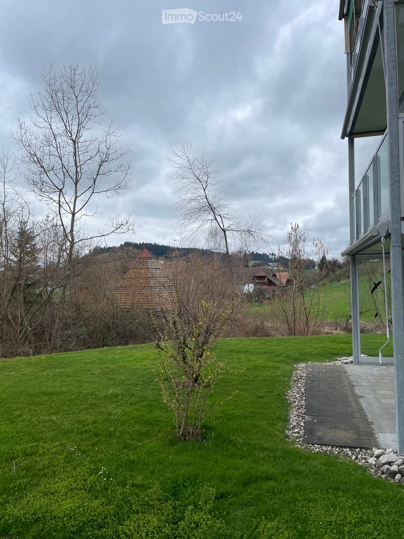 Lush green lawn with a small tree, a pathway leading to a modern house with large windows, and a view of a rural landscape with hills and traditional houses in the background.