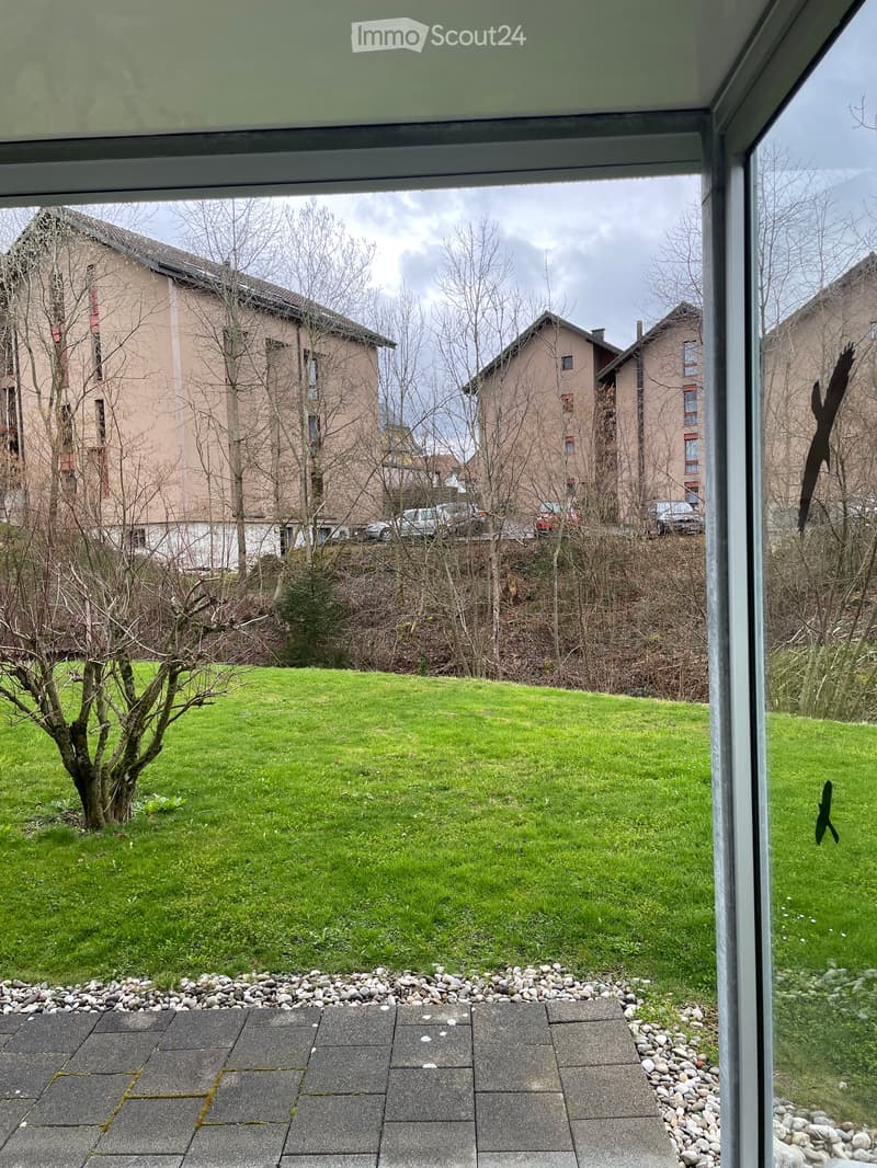 Balcony with a view of a green lawn, small trees, and neighboring buildings in the background.