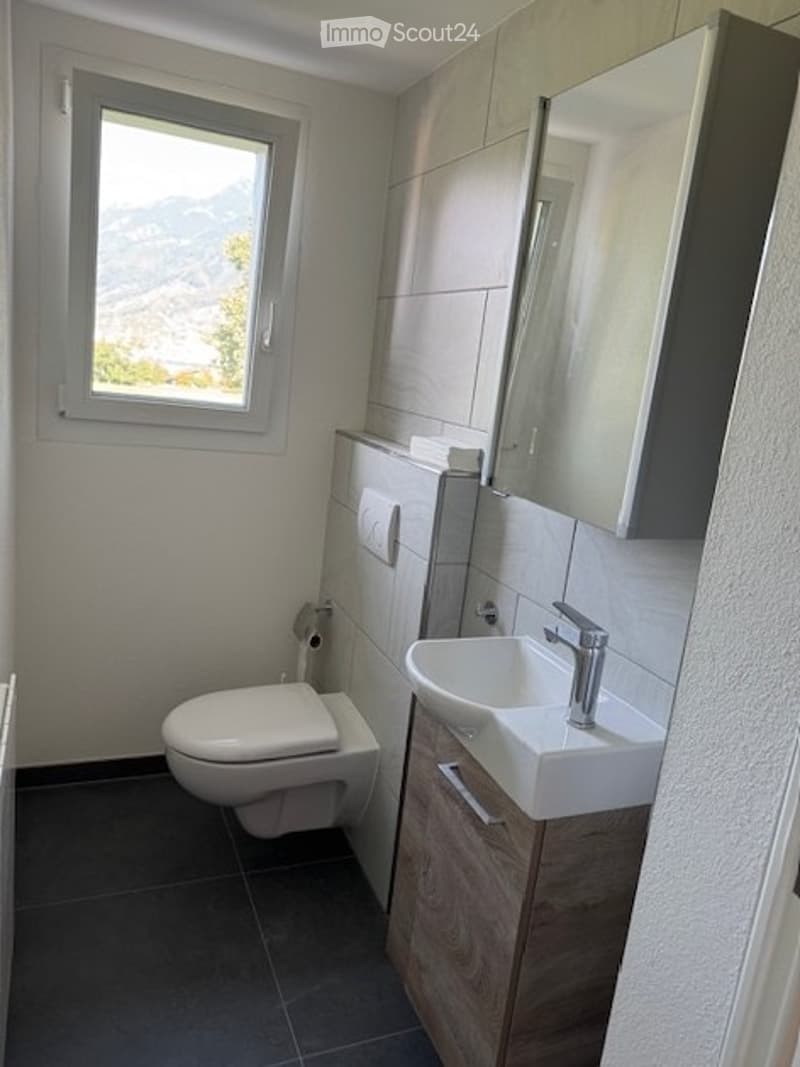 Modern bathroom with a toilet, sink, mirror, and cabinet. The room features white tiles, a window with a scenic view, and dark flooring.