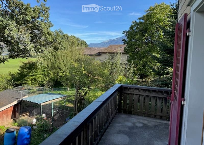 Balcony overlooking a lush garden and distant mountains, with wooden railing and a view of the surrounding landscape.