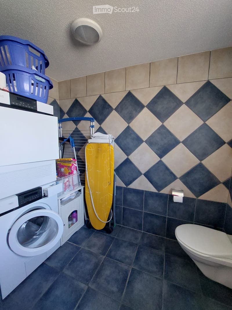 Bathroom with washing machine, ironing board, toilet, laundry basket, and shelf.