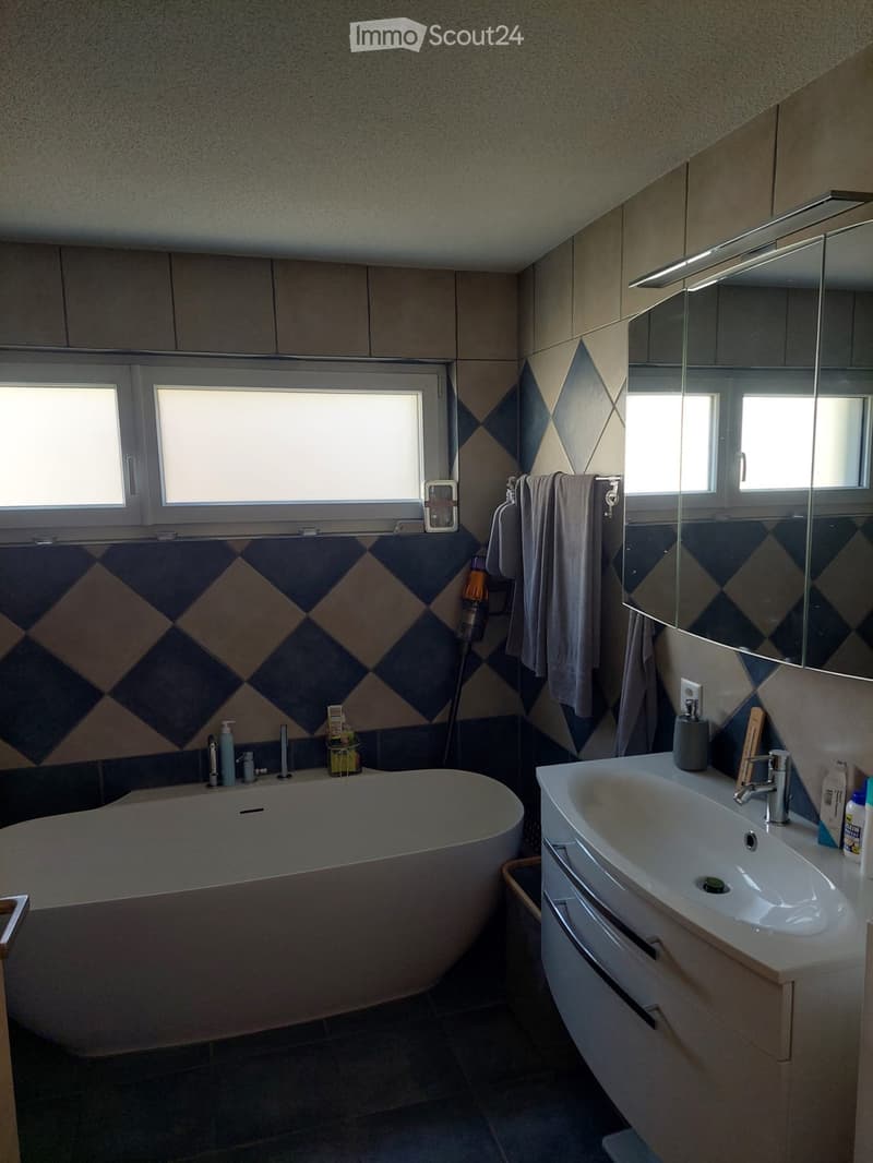 Modern bathroom with a bathtub, sink, and large mirror. Features diamond-patterned tiles and natural light from windows.