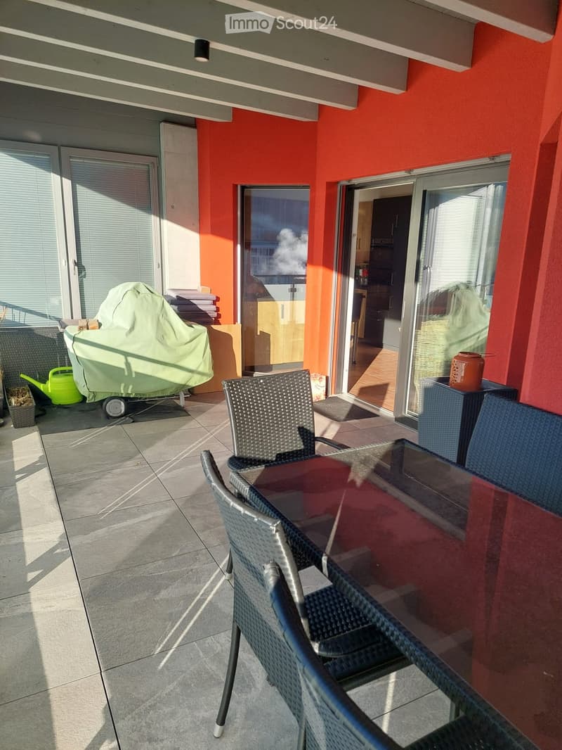 Balcony with tiled floor, glass table, wicker chairs, covered grill, and a view of the city.