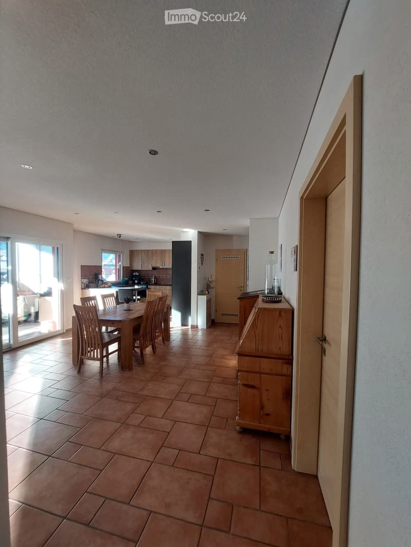 Open-plan dining and kitchen area with tiled flooring, wooden dining table and chairs, kitchen cabinets, and a bottle on a sideboard.